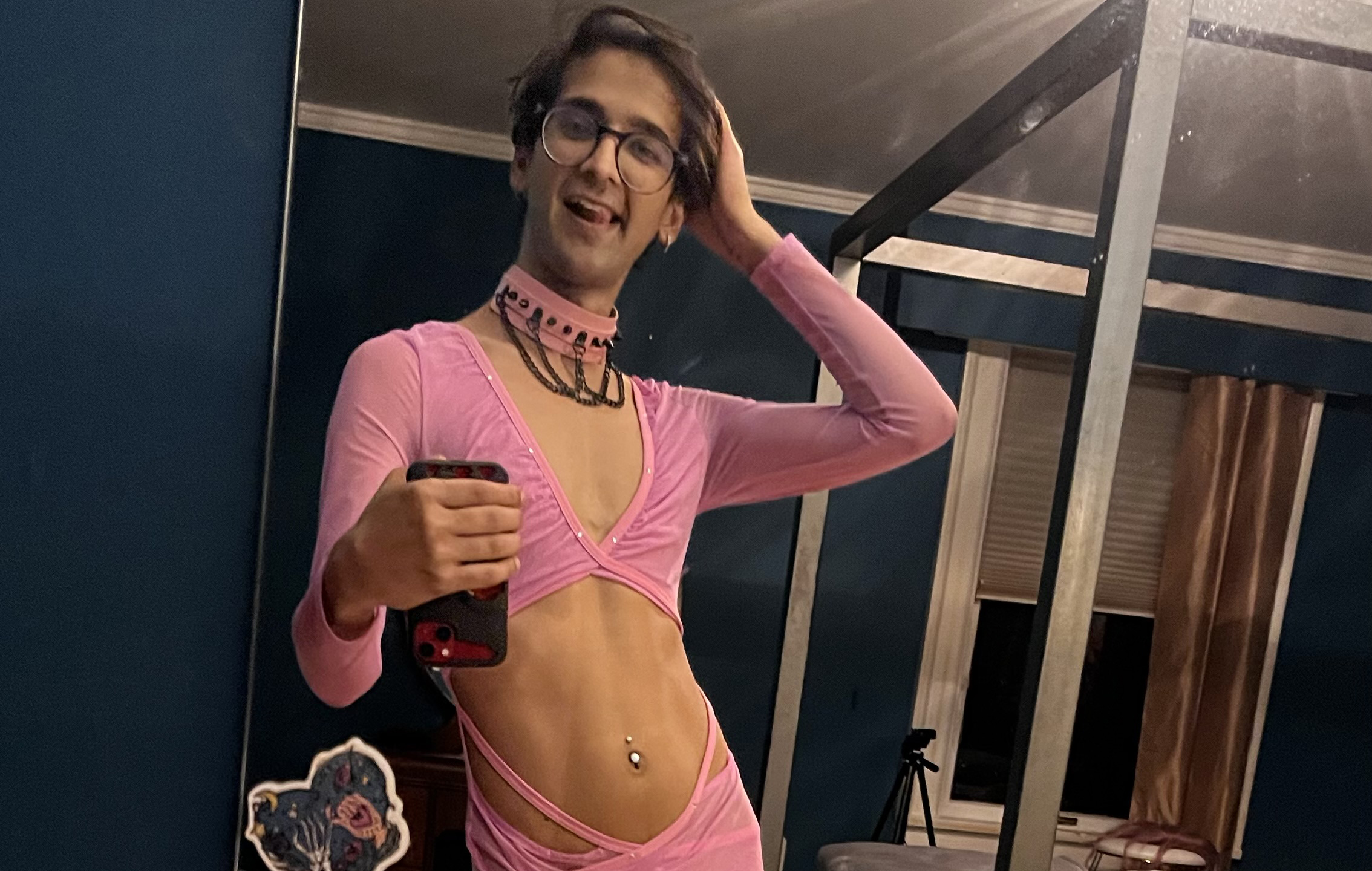 A smiling femboy in a sheer pink top and a studded choker taking a mirror selfie