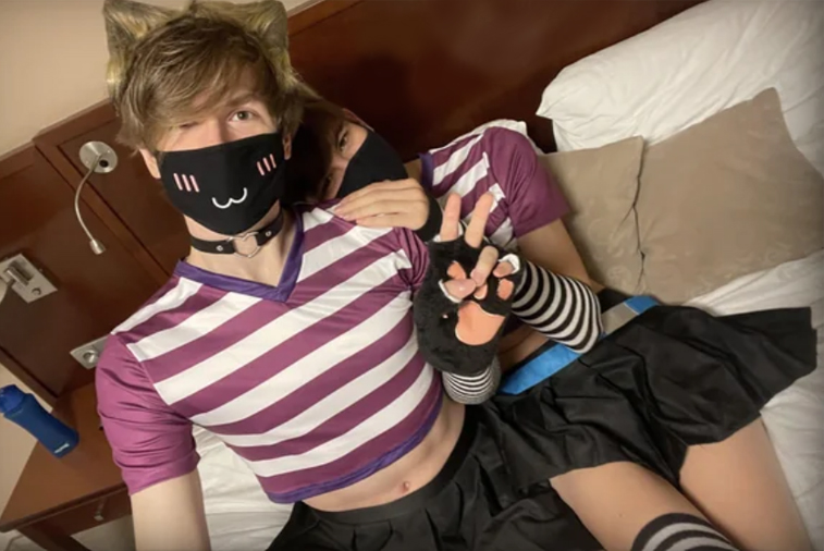 A femboy in a striped crop top, pleated skirt, thigh-high socks and a cat-ear headband lying on a bed and making a peace sign