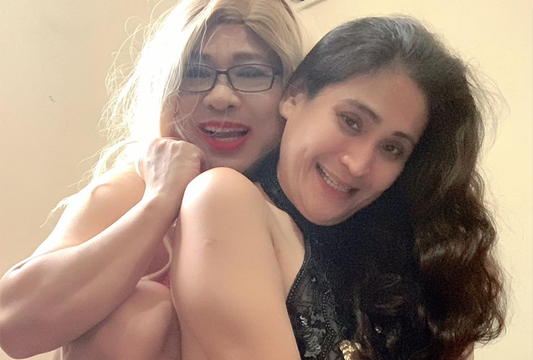 Two people cheek to cheek in a selfie, both laughing — one with blonde hair and glasses, one with long dark hair